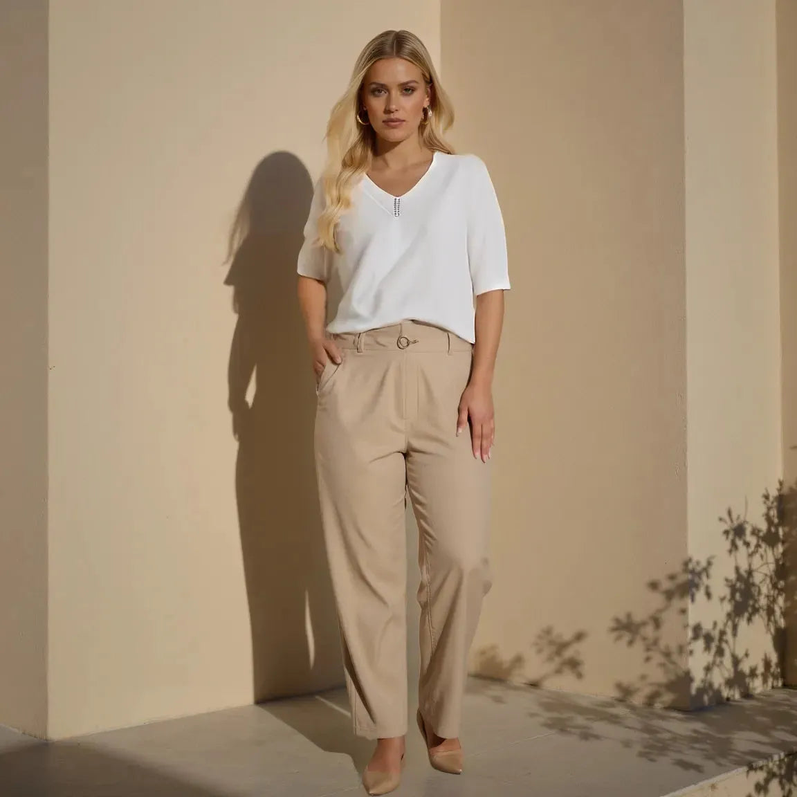 Woman wearing a white top and beige pants against a beige wall.