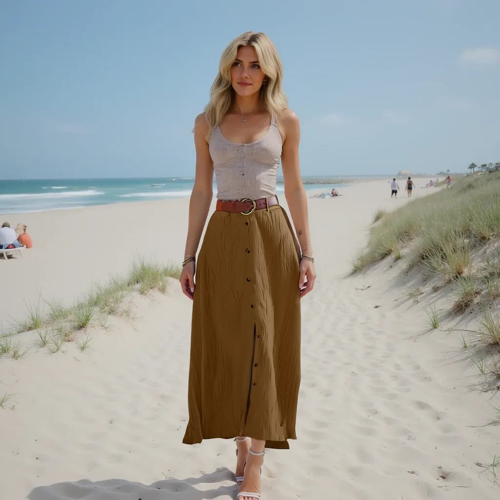 Woman standing on a sandy beach wearing a white top and brown skirt.
