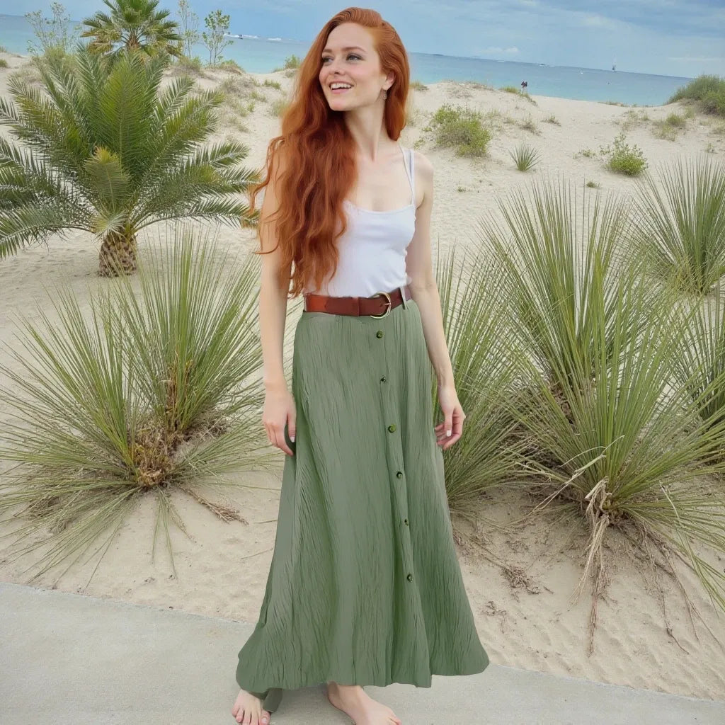 Woman in a green skirt and white top standing on a sandy beach with palm trees and ocean in the background.
