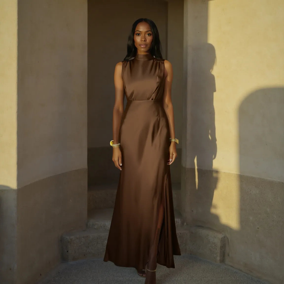 Woman in a brown dress standing in a sunlit room with shadows on the wall.