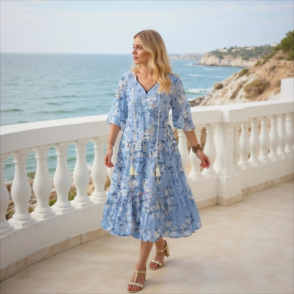 Woman in a blue floral dress standing on a balcony overlooking the ocean.