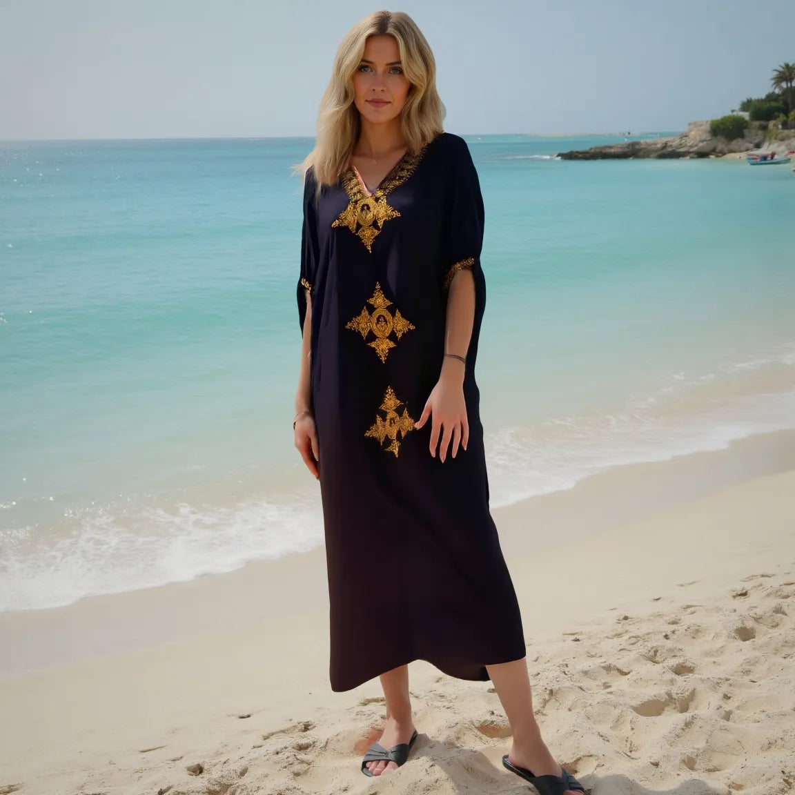 Woman in a black dress with gold patterns standing on a beach.