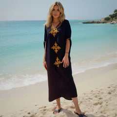 Woman in a black dress with gold patterns standing on a beach.