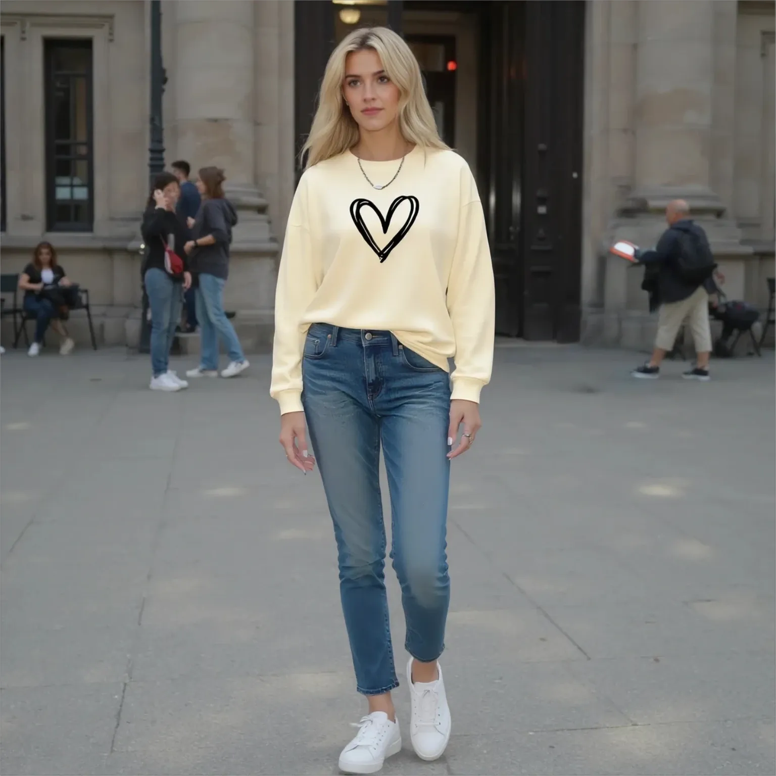 Woman wearing a beige sweatshirt with a heart design, blue jeans, and white sneakers on a city street.