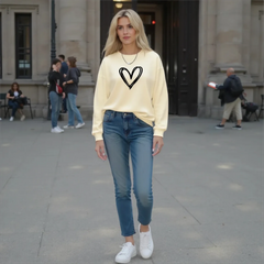 Woman wearing a beige sweatshirt with a heart design, blue jeans, and white sneakers on a city street.