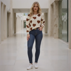 Woman wearing a cream sweater with brown heart patterns and dark blue jeans in a hallway.