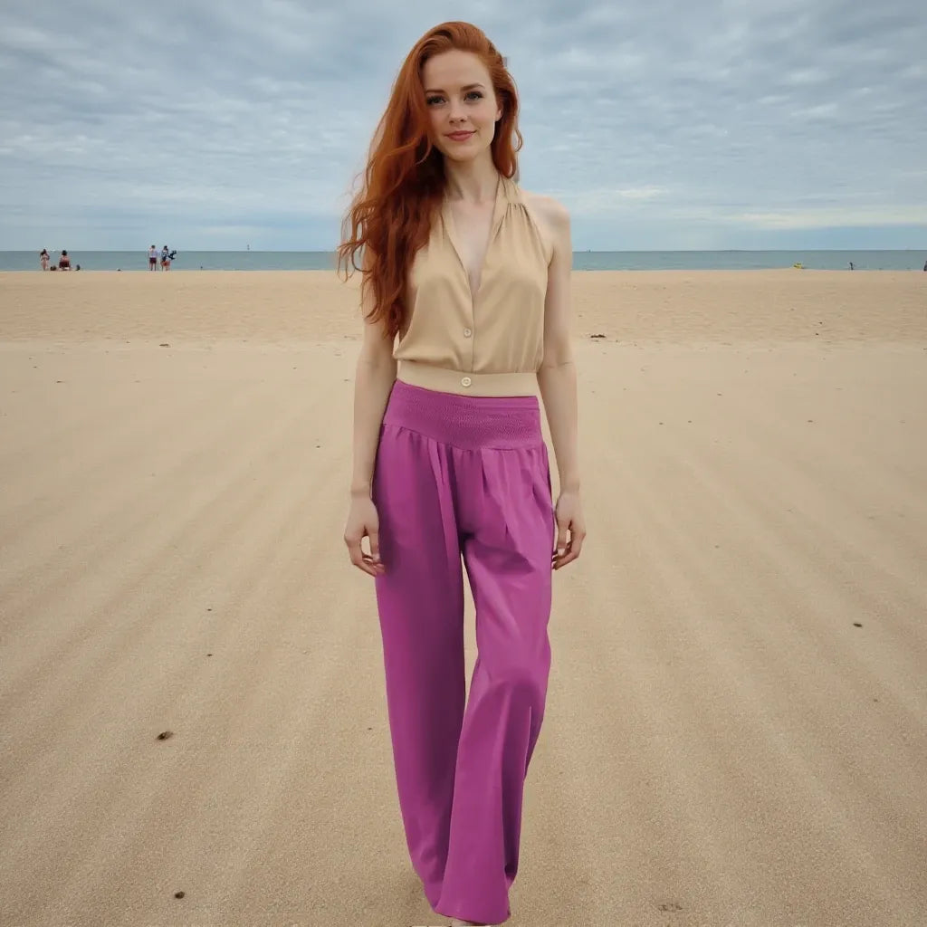 Woman in beige top and pink pants standing on a sandy beach with ocean in the background