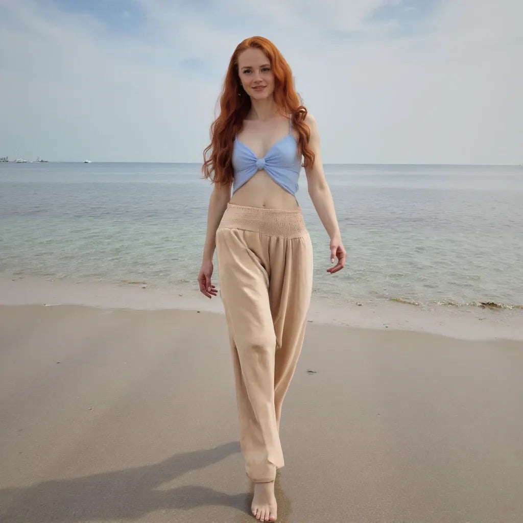 Woman standing on a beach wearing a blue top and beige pants with ocean in the background
