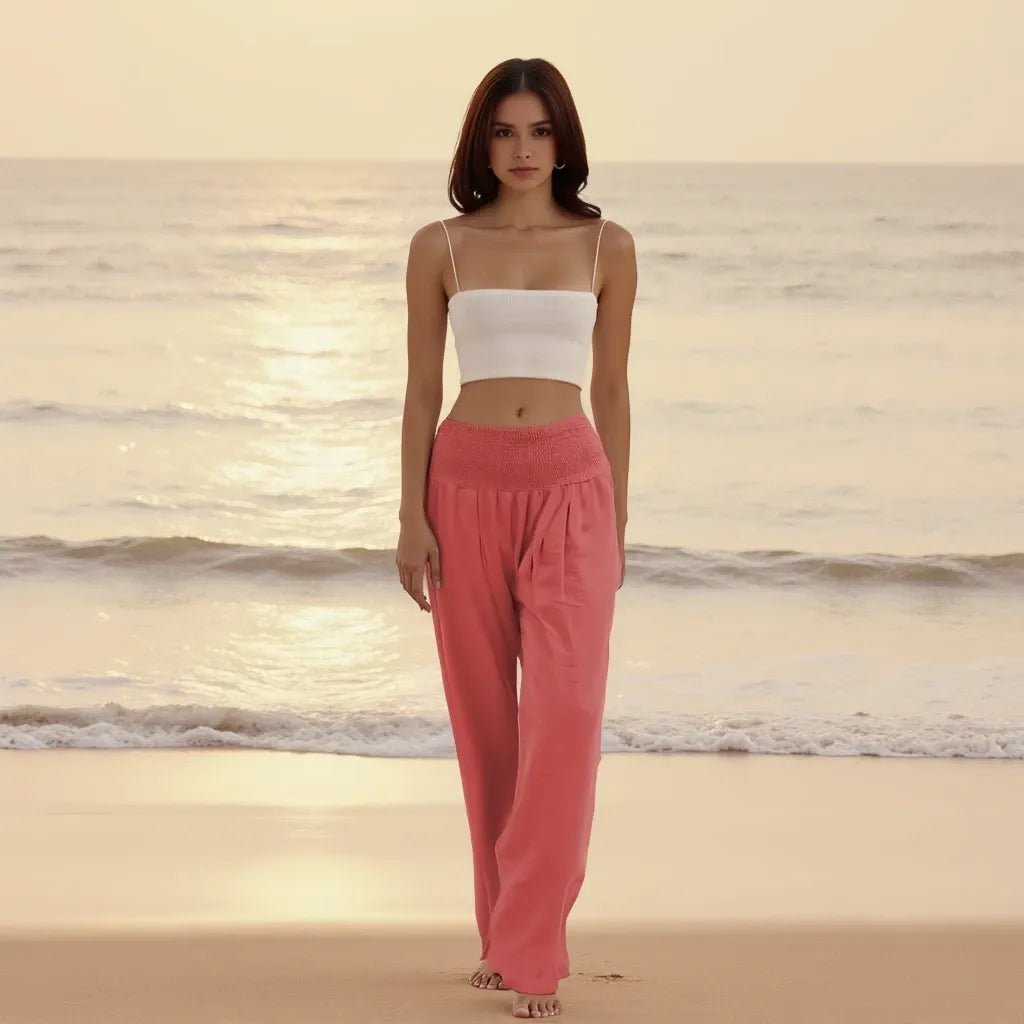 Woman wearing a white top and pink pants standing on a beach with ocean waves in the background.
