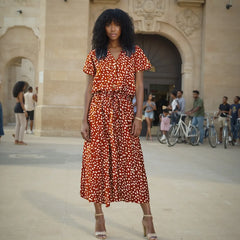 Woman in a red patterned dress standing in an outdoor setting with people and bicycles in the background.