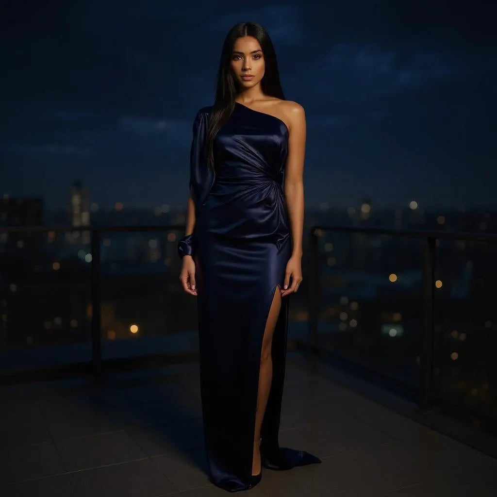 Woman in a dark blue one-shoulder gown standing on a rooftop at night with city lights in the background.