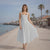 Woman in a white dress standing on a beach promenade with palm trees and buildings in the background.