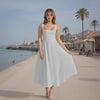 Woman in a white dress standing on a beach promenade with palm trees and buildings in the background.