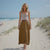 Woman standing on a sandy beach wearing a white top and brown skirt.