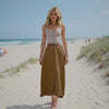 Woman standing on a sandy beach wearing a white top and brown skirt.