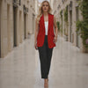 Woman in a red blazer and black pants walking down a sunlit street.