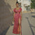 Woman in a red patterned dress standing on a street with a stone wall and building in the background.