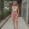 Woman in a white top and pink shorts standing on a sidewalk.