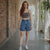 Woman wearing a brown tank top and blue denim shorts standing in a sunlit hallway with brick walls and plants.