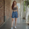 Woman wearing a brown tank top and blue denim shorts standing in a sunlit hallway with brick walls and plants.