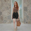 Woman with red hair standing in front of a stone wall holding a brown handbag.