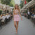 Woman in a pink outfit walking on a sidewalk with outdoor dining tables in the background