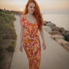 Woman in a floral dress standing on a path by a beach at sunset.