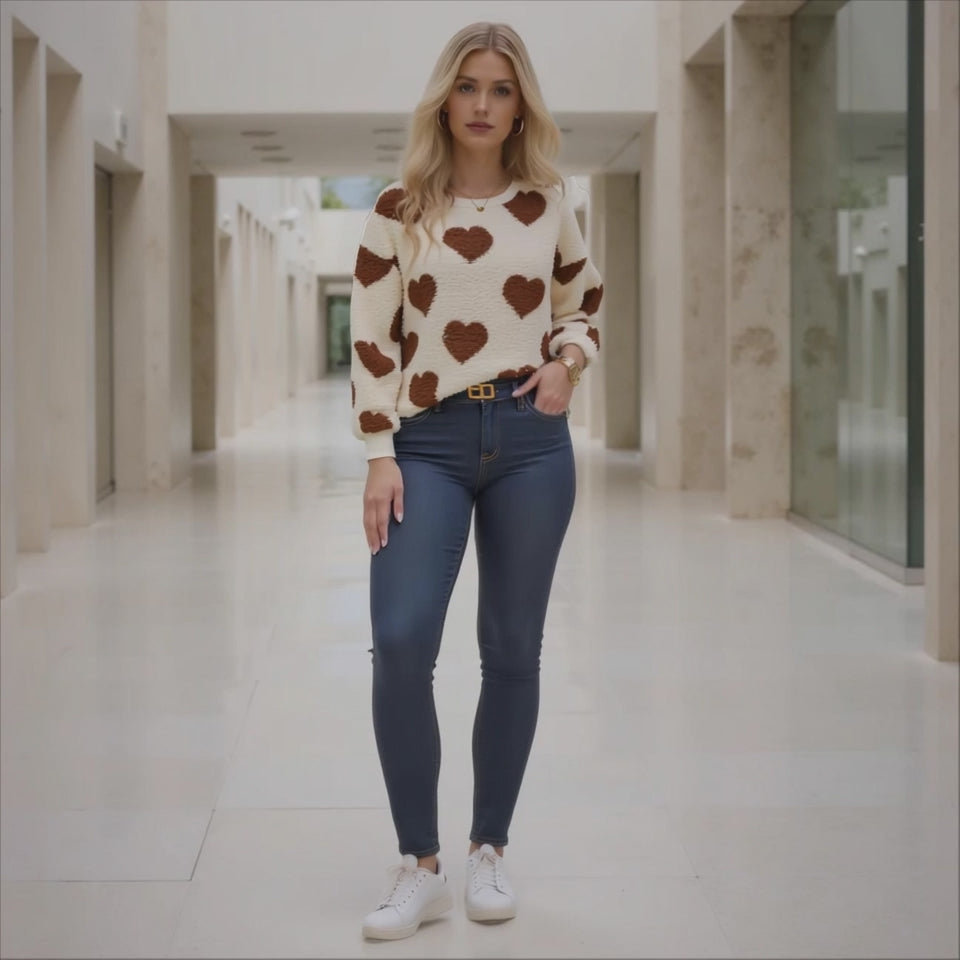 Woman wearing a cream sweater with brown heart patterns and dark blue jeans in a hallway.