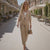 Woman in a beige outfit walking on a sunlit street with cafes and greenery.