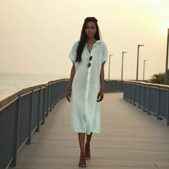 Woman in a light green dress standing on a wooden boardwalk by the water.