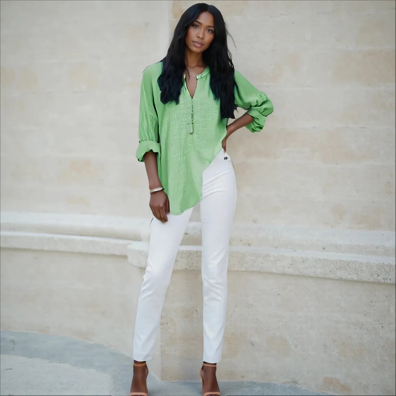 Woman wearing a green shirt and white pants standing against a beige wall.