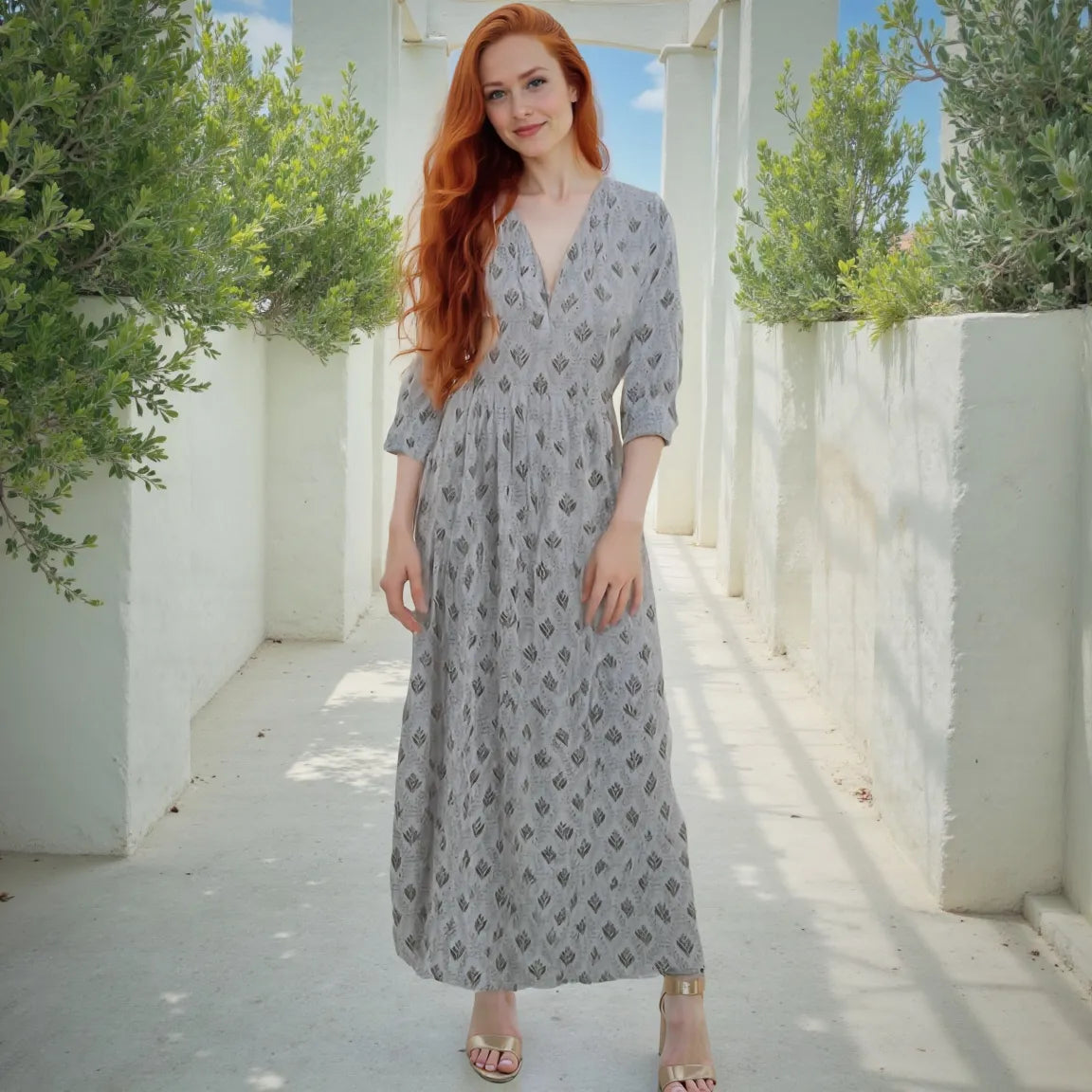 Woman in a gray patterned dress standing in a sunlit outdoor setting with white columns and greenery.