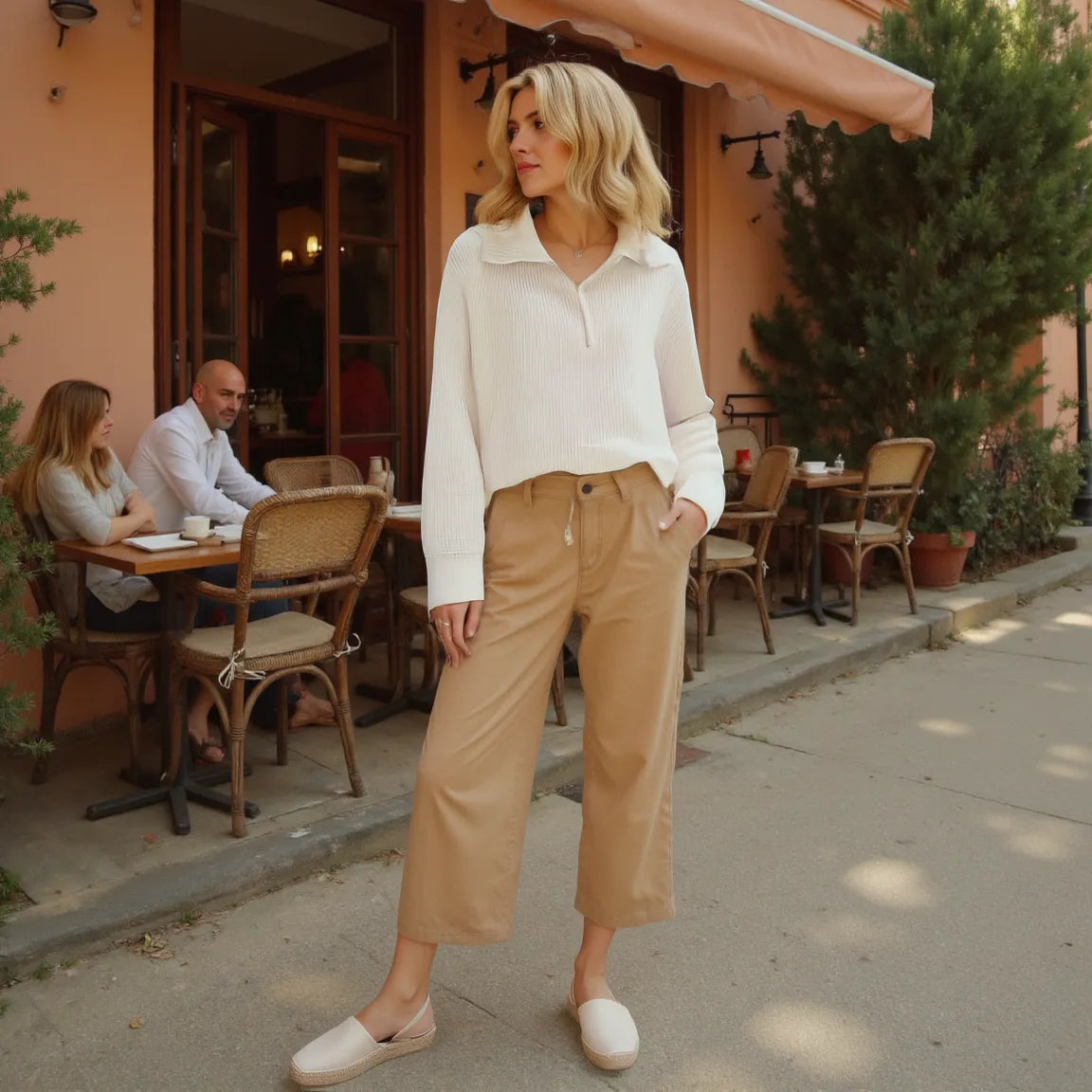 Woman in a white blouse and beige pants standing on a sidewalk with a cafe in the background