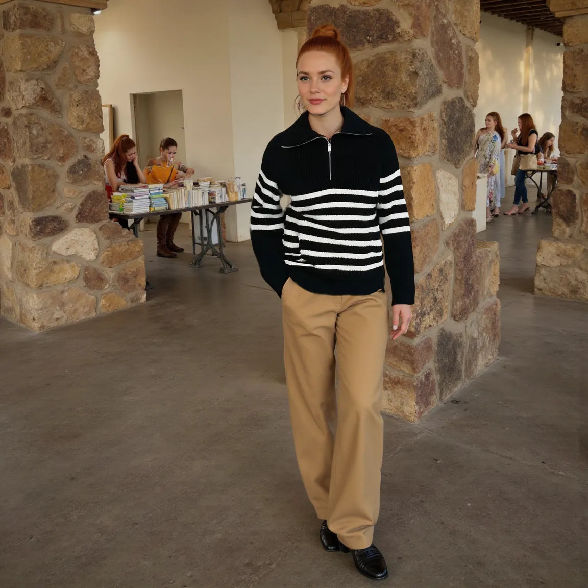 Woman in a black and white striped sweater and beige pants standing in a stone-walled room with people in the background.