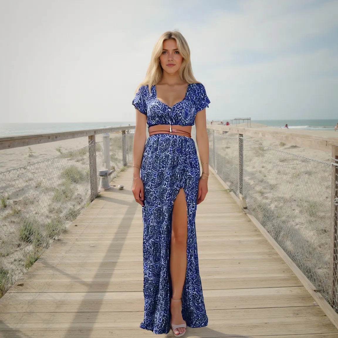 Woman in a blue patterned dress standing on a wooden boardwalk by the beach.