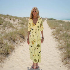 Woman in a yellow floral dress walking on a sandy path with grasses and a clear sky.
