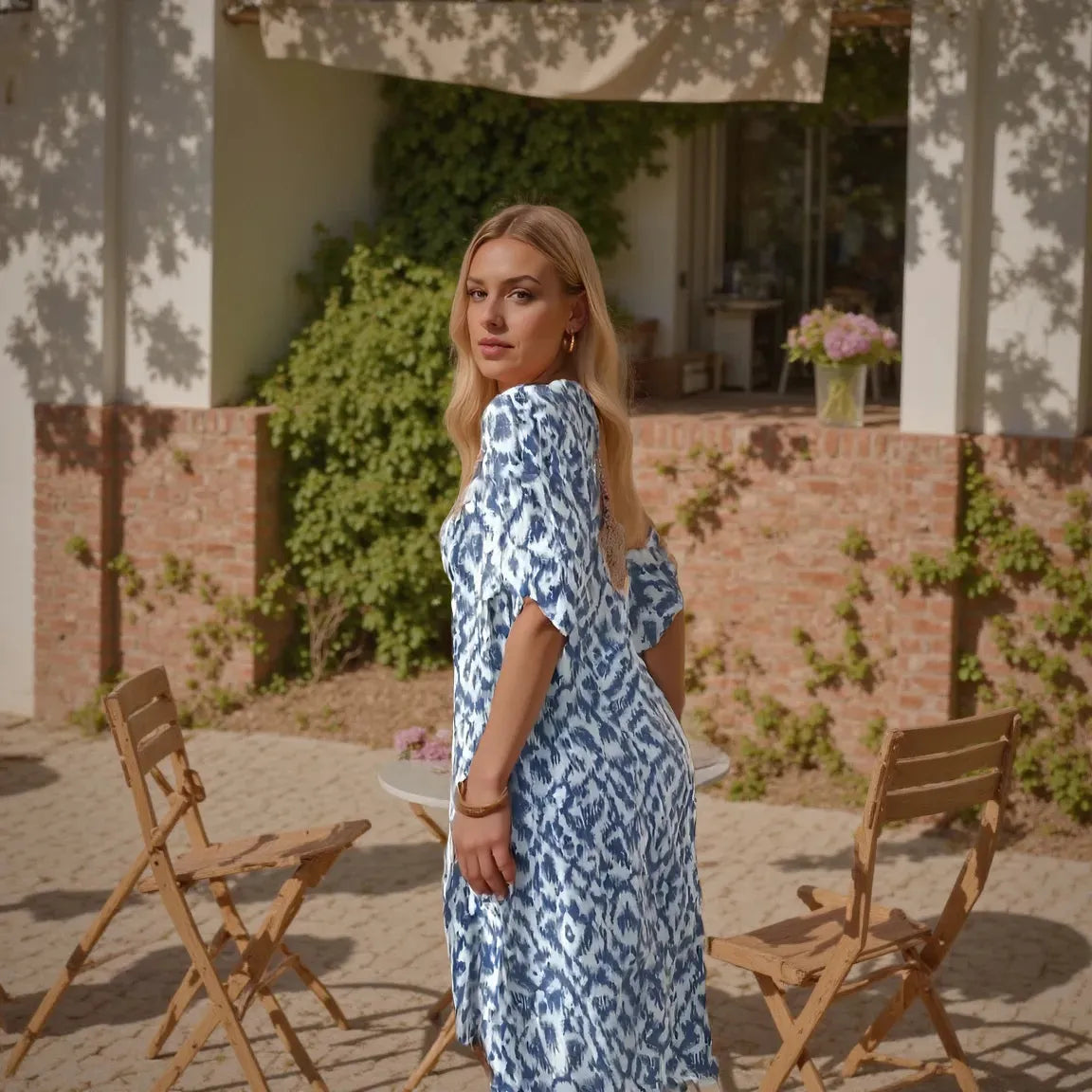 Woman in a blue patterned dress standing outdoors near wooden chairs and a table.