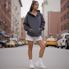 Woman in a gray hoodie and white shorts standing on a city street with cars and buildings in the background.