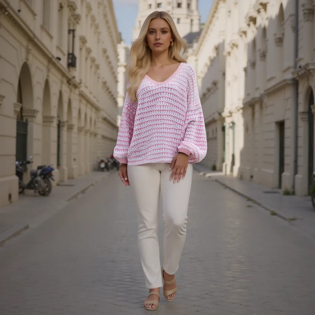 Woman wearing a pink knitted sweater and white pants walking down a sunlit street.