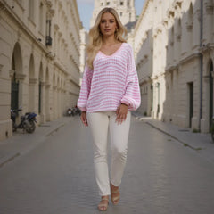 Woman wearing a pink knitted sweater and white pants walking down a sunlit street.
