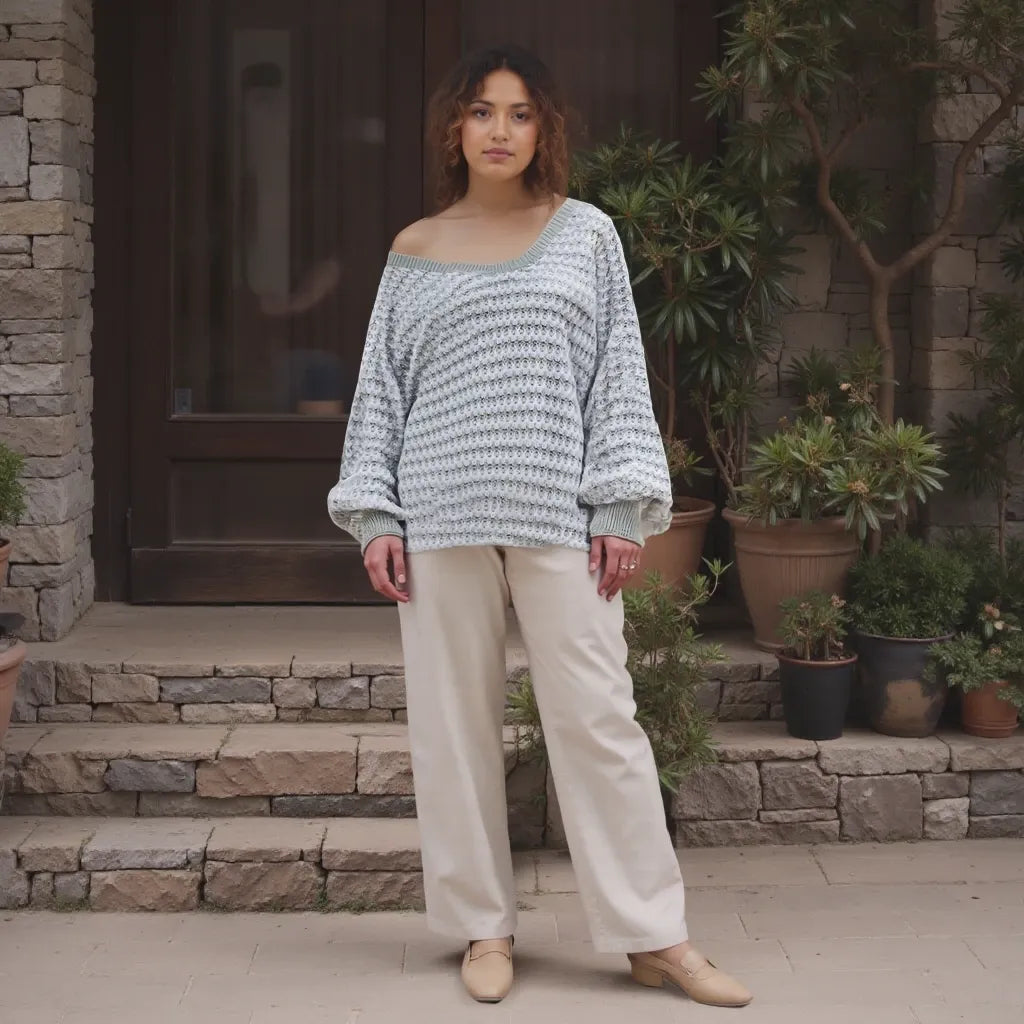 Woman wearing a light blue patterned sweater and beige pants standing in front of a stone building with plants.