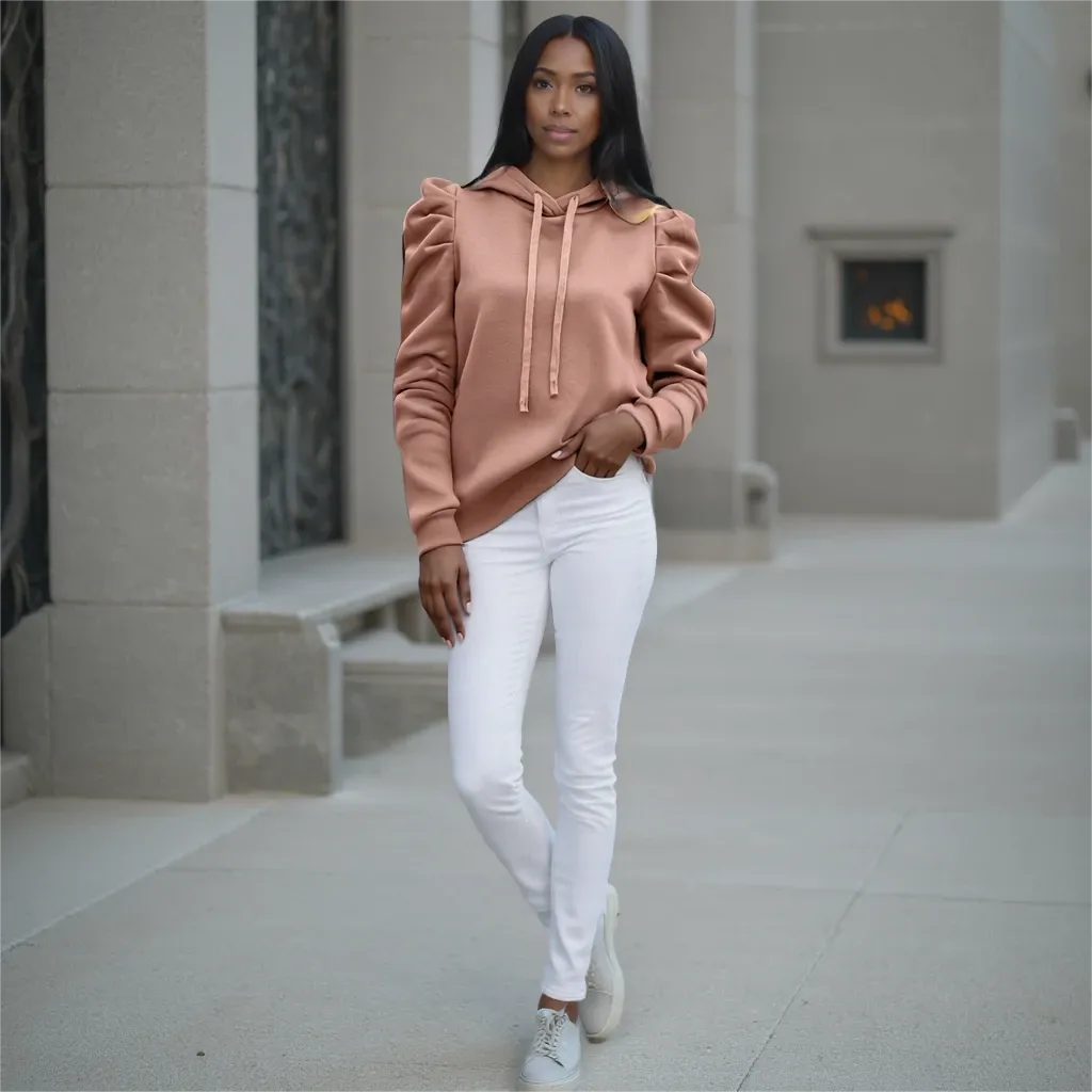 Woman wearing a peach hoodie and white pants standing in an indoor setting.