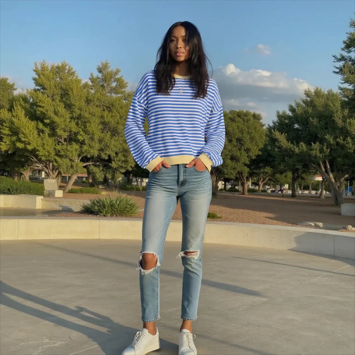 Woman wearing a blue and white striped sweater and ripped jeans standing outdoors with trees in the background.