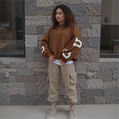 Woman wearing a brown sweater with white lettering and beige pants standing against a stone wall.